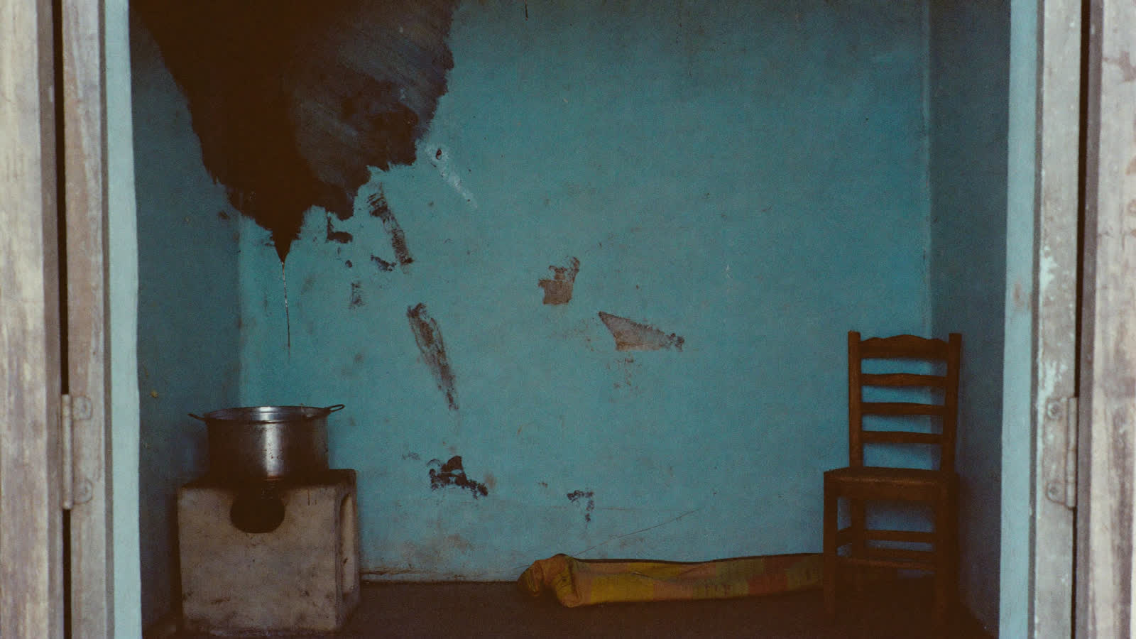 A bare room with a monsoon stain bleeding down the wall, one pot on a small stove, and an empty chair.